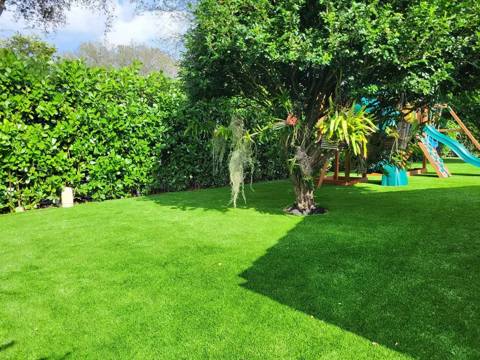 A sunny backyard features a vibrant green lawn with a tree at the center. To the right, a children's playset with a slide stands proudly. Lush hedges border the area, creating seclusion under the clear blue sky above, while synthetic grass ensures year-round perfection and easy maintenance.