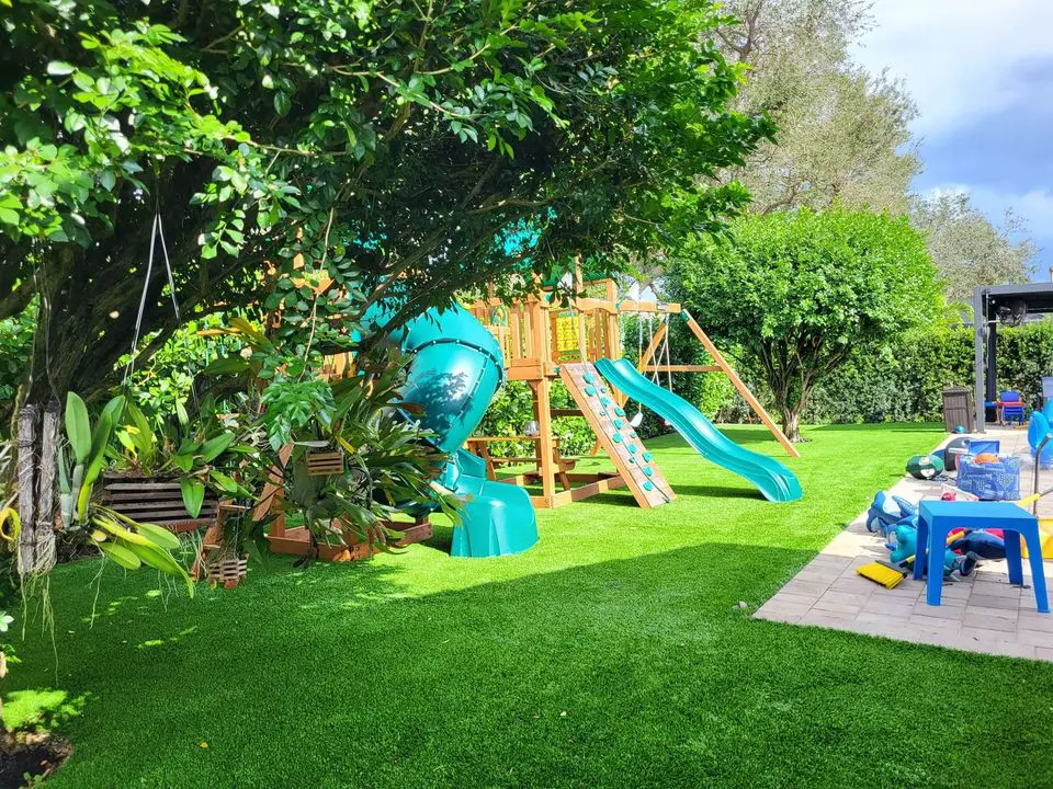 A backyard playground in Surprise AZ features a wooden playset with two green slides and a climbing wall. A blue spiral slide peeks through lush greenery. The area, enhanced by artificial grass, includes a paved patio with a blue chair and scattered toys on the lawn.