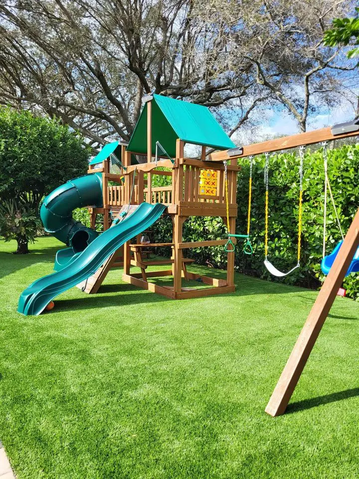 A backyard playground set with a green slide, tunnel, and swings nestles in lush greenery and trees. The wooden frame boasts a green canopy over a lawn of synthetic turf. Above, the sky remains blue with a few clouds.