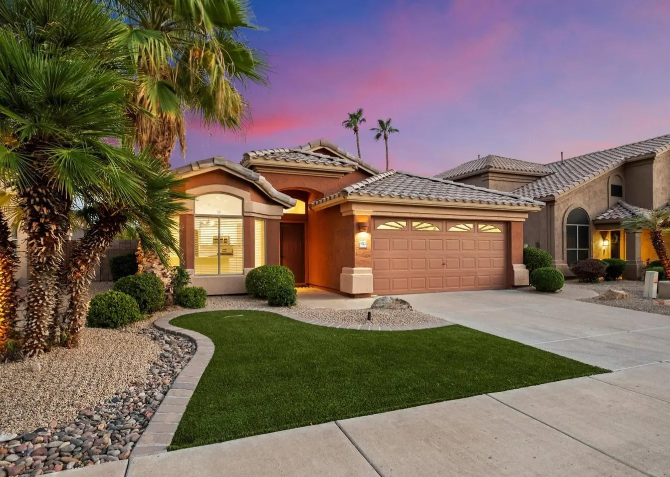 Single-story house in El Mirage, AZ, featuring a two-car garage. Bordered by palm trees and desert landscaping with artificial turf installation, the tan exterior and tiled roof complement the sunset's pink and purple hues in the sky.