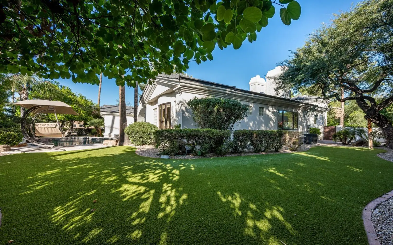A well-maintained backyard in, Surprise, AZ, features a lush green lawn bordered by shrubs and trees. A comfortable seating area with a canopy is visible near the left, and the house is partially visible on the right. The scene is sunlit with a clear blue sky.