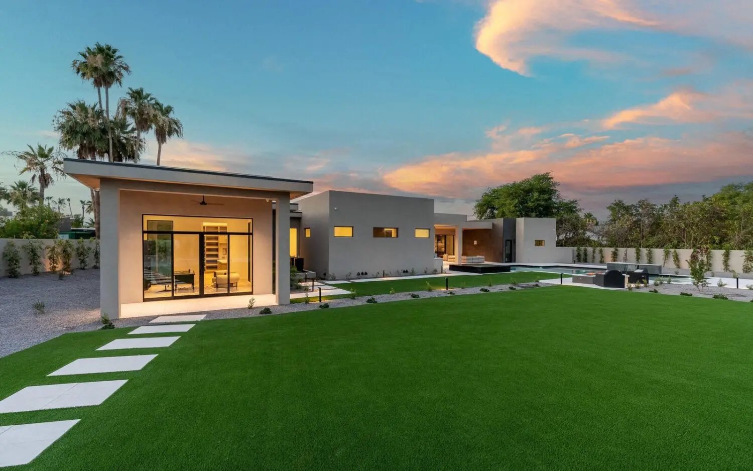 Modern house with large glass windows and a flat roof, surrounded by a spacious lawn featuring synthetic turf and stepping stones. The sky is partly cloudy with a mix of pink and blue hues, and there are palm trees and shrubs in the background.