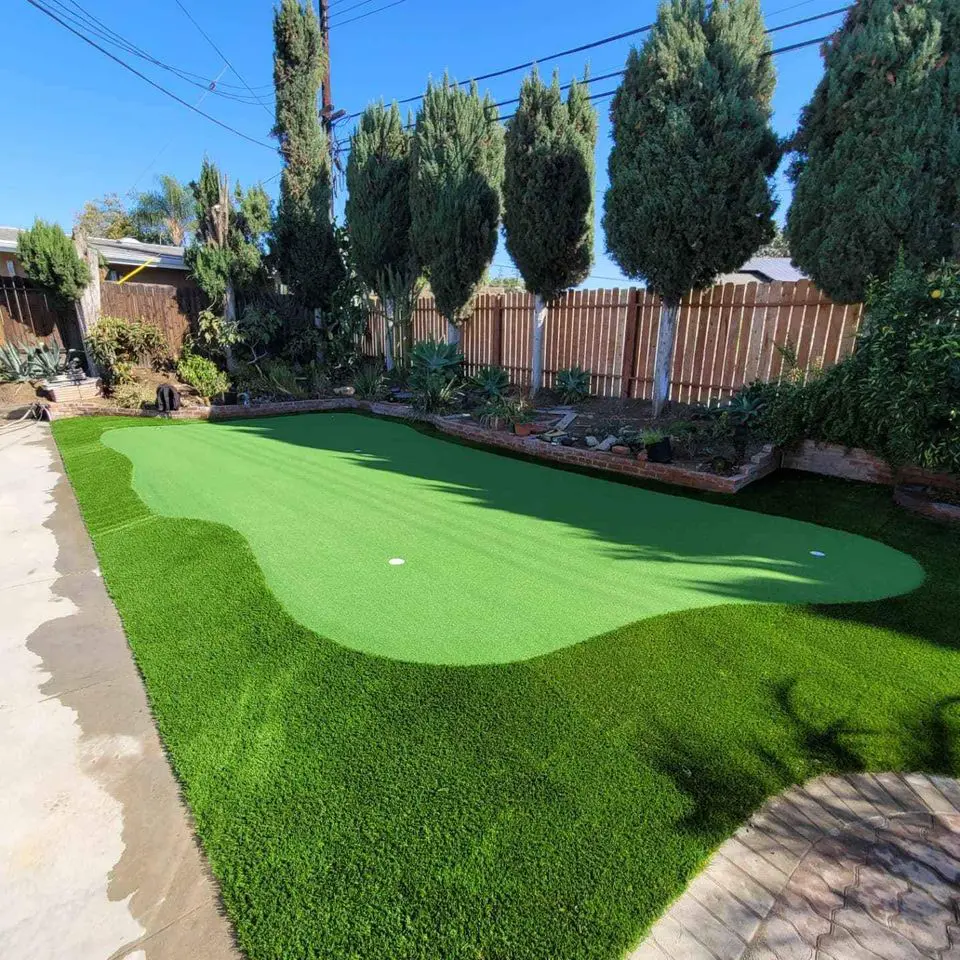 A backyard featuring a small putting green with two white holes, surrounded by lush synthetic grass and bordered by tall trees and a wooden fence. This low-maintenance lawn flourishes under the clear, blue sky above.