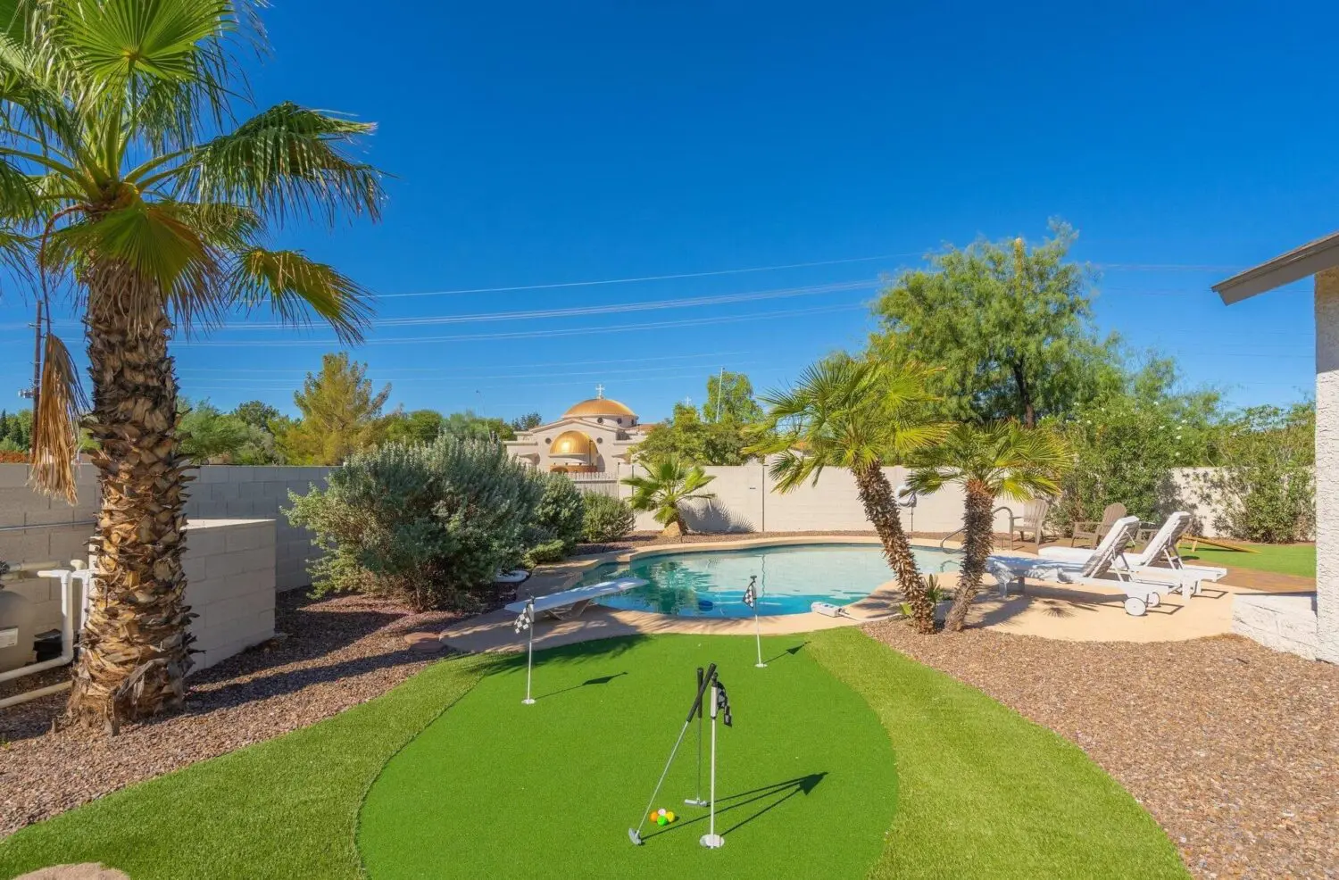 In Surprise, AZ, a backyard oasis features a small putting green with pristine artificial grass amid tropical plants and a shimmering pool. Lounge chairs beckon by the water's edge under a clear blue sky, and in the background, a charming dome-shaped building completes this serene escape.