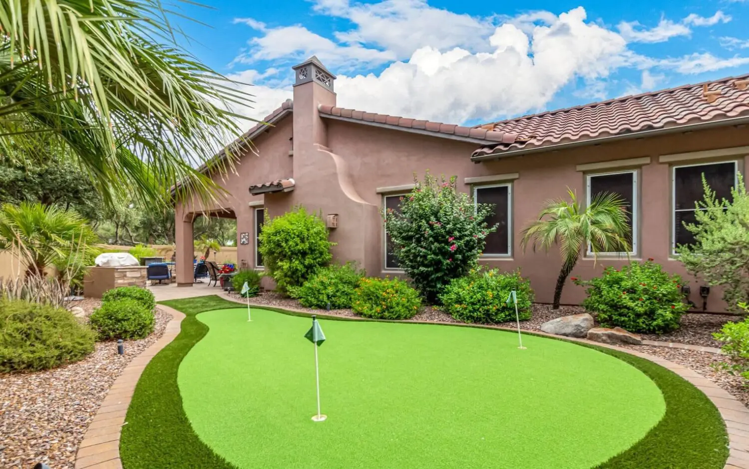 A backyard in Peoria, AZ features a small putting green with golf flags, surrounded by a well-maintained garden of bushes and palm trees. The synthetic turf adds vibrancy, while the house with its terracotta roof and stucco walls sits under a partly cloudy blue sky.