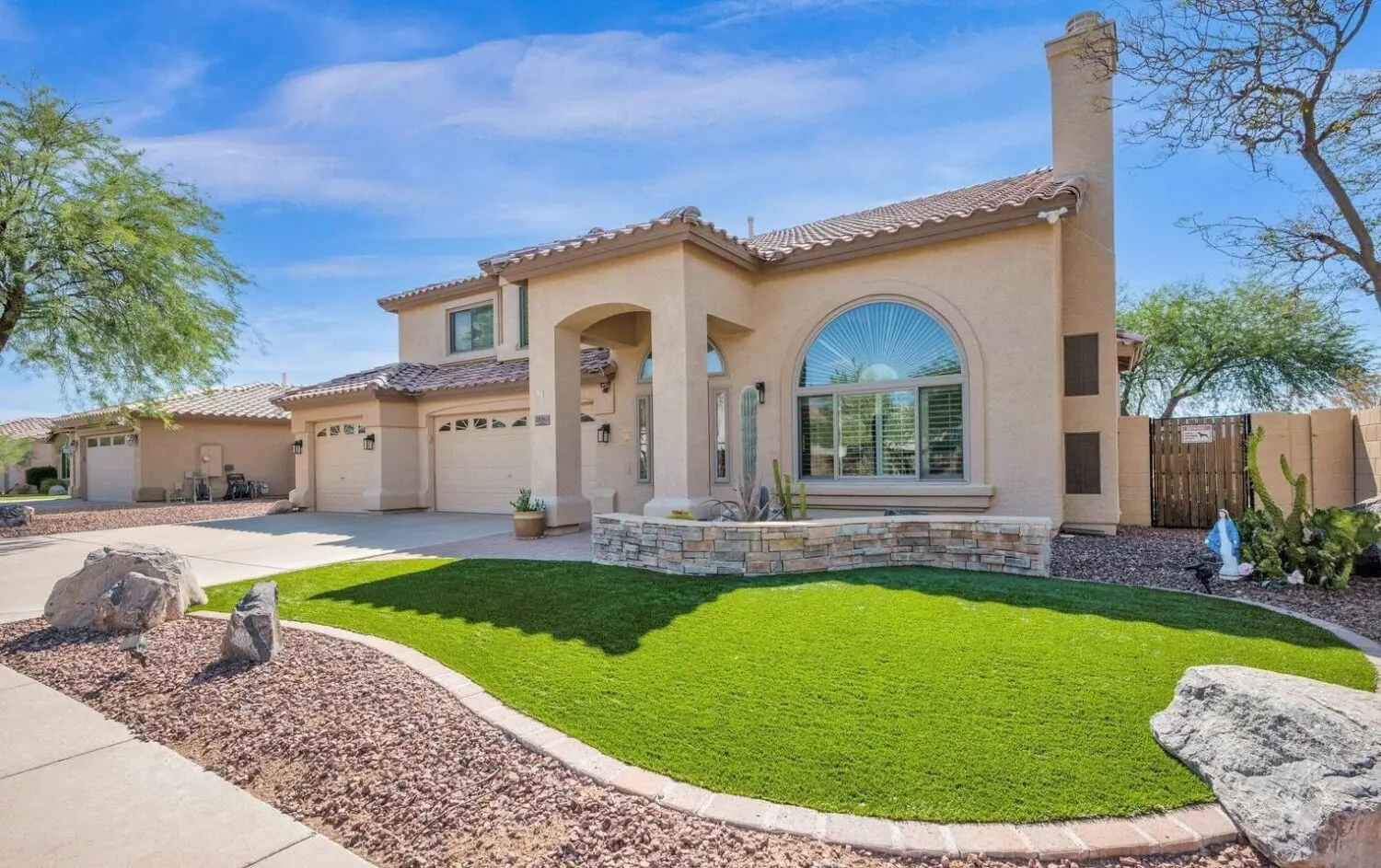 A modern two-story house in Surprise, AZ, with a stucco exterior and tiled roof. It features a large arched window, artificial grass on the well-maintained front lawn, stone accents, and desert landscaping. The sky is clear and blue.