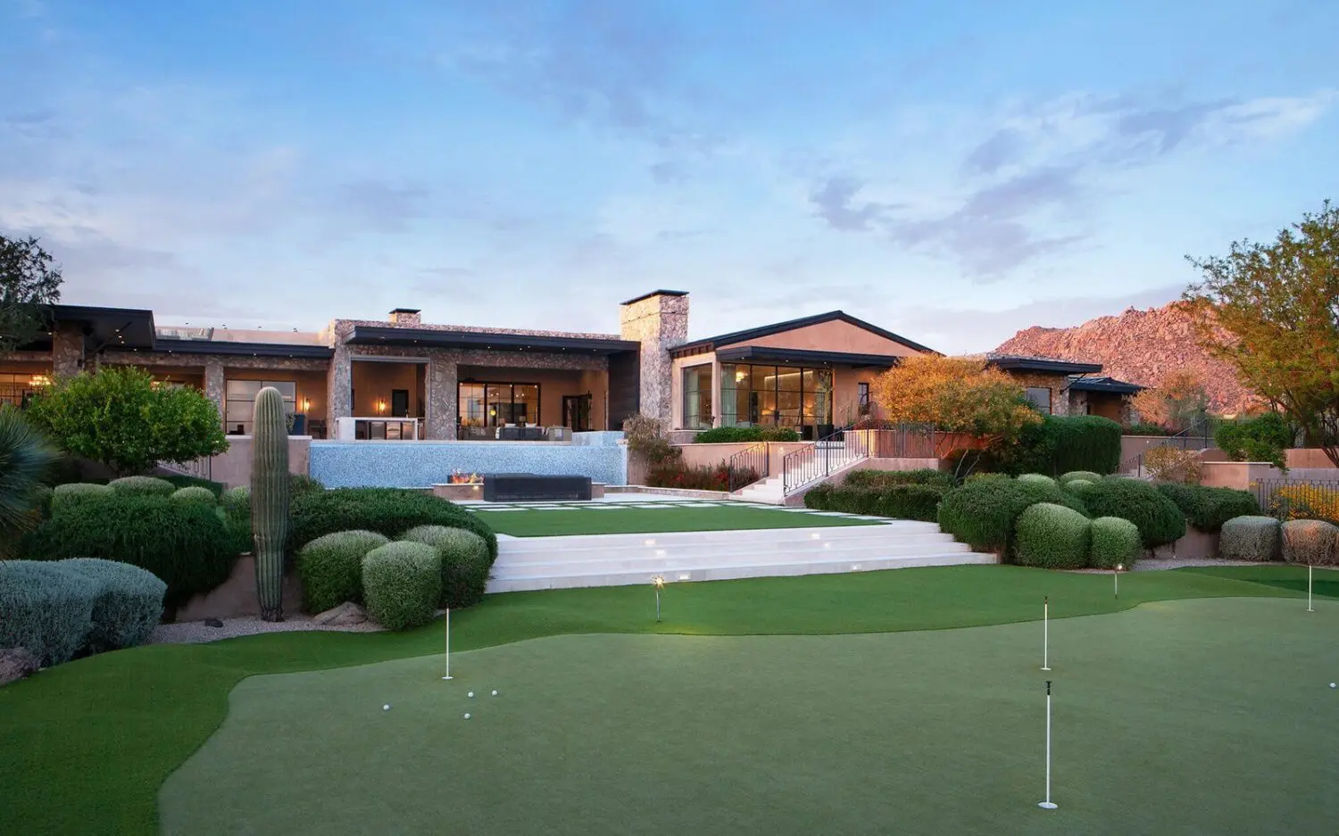 Luxury modern house with large windows, surrounded by manicured landscaping and a putting green featuring artificial grass in the foreground. The setting includes a mountain view under a clear blue sky.