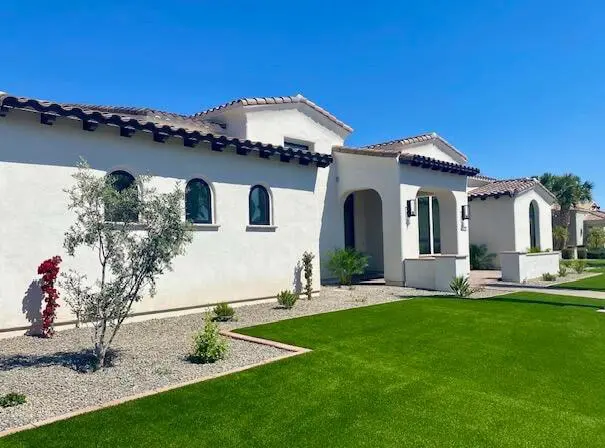 A modern, single-story white stucco house in El Mirage, AZ, showcases arched windows and a clay tile roof. The neatly landscaped front yard features green grass and sparse desert plants under a clear blue sky, highlighting its ideal blend of residential and commercial appeal.