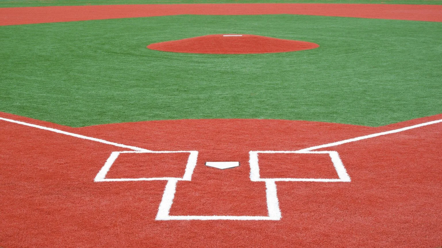 A baseball infield with a focus on home plate in the foreground and the pitcher's mound in the background. The field features a red clay color for the base paths and vibrant synthetic grass for the playing area, offering a low-maintenance lawn that perfectly complements its rich hues.