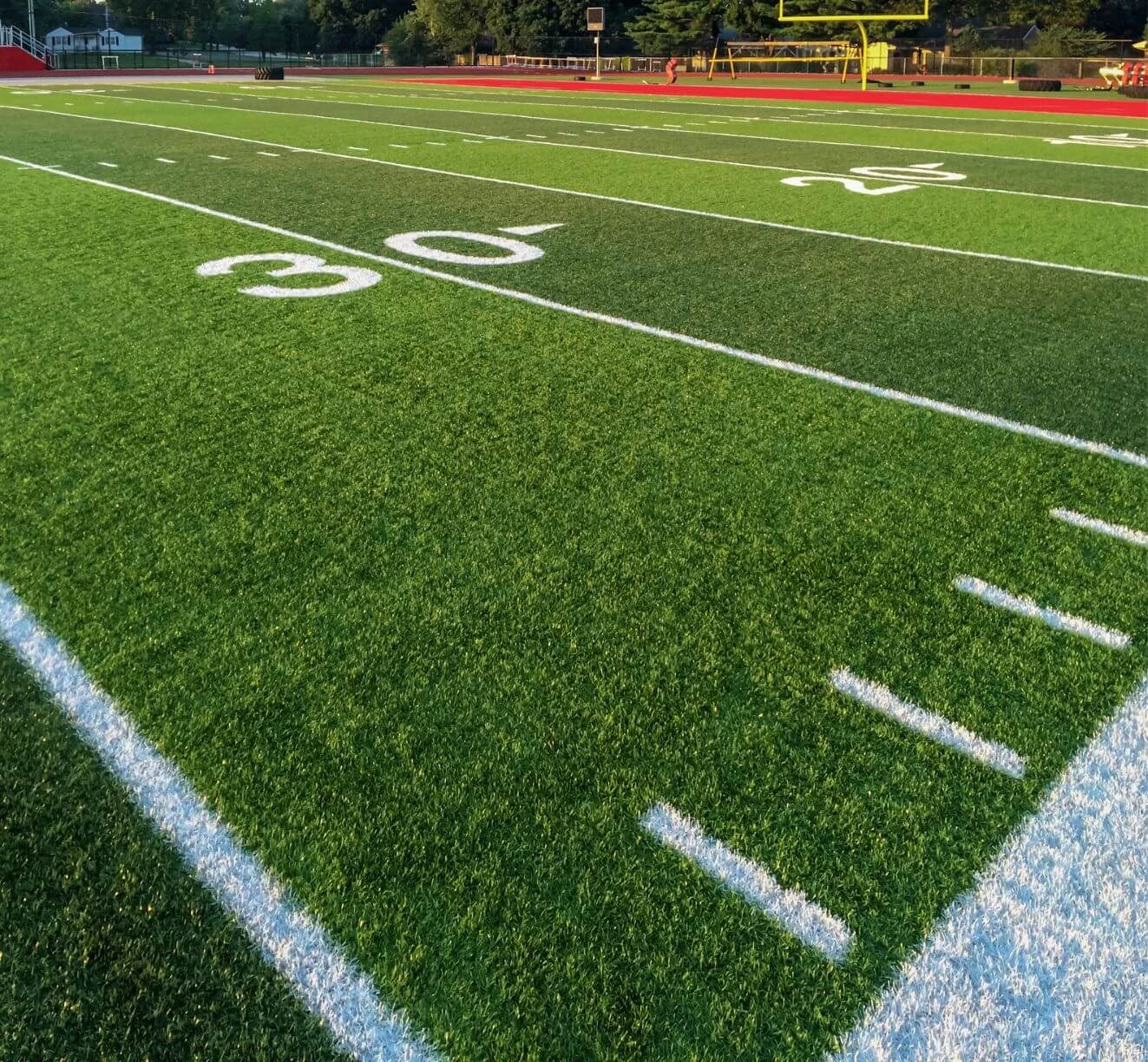 A football field with bright green artificial turf and white yard lines, viewed from the 30-yard line. The field has a visible track around it and a goalpost at the far end. Lush trees are in the background of El Mirage, AZ, under a clear sky.