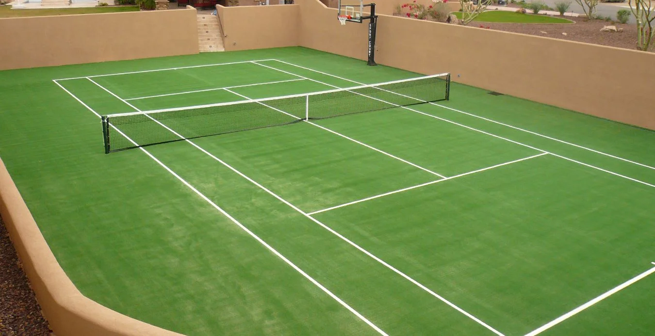 An outdoor sports court featuring an artificial turf installation with a green surface and white lines, resembling a tennis court with a basketball hoop on one end. It is enclosed by tan walls and has a small staircase leading up from the court area, perfect for residential and commercial use in Surprise, AZ.