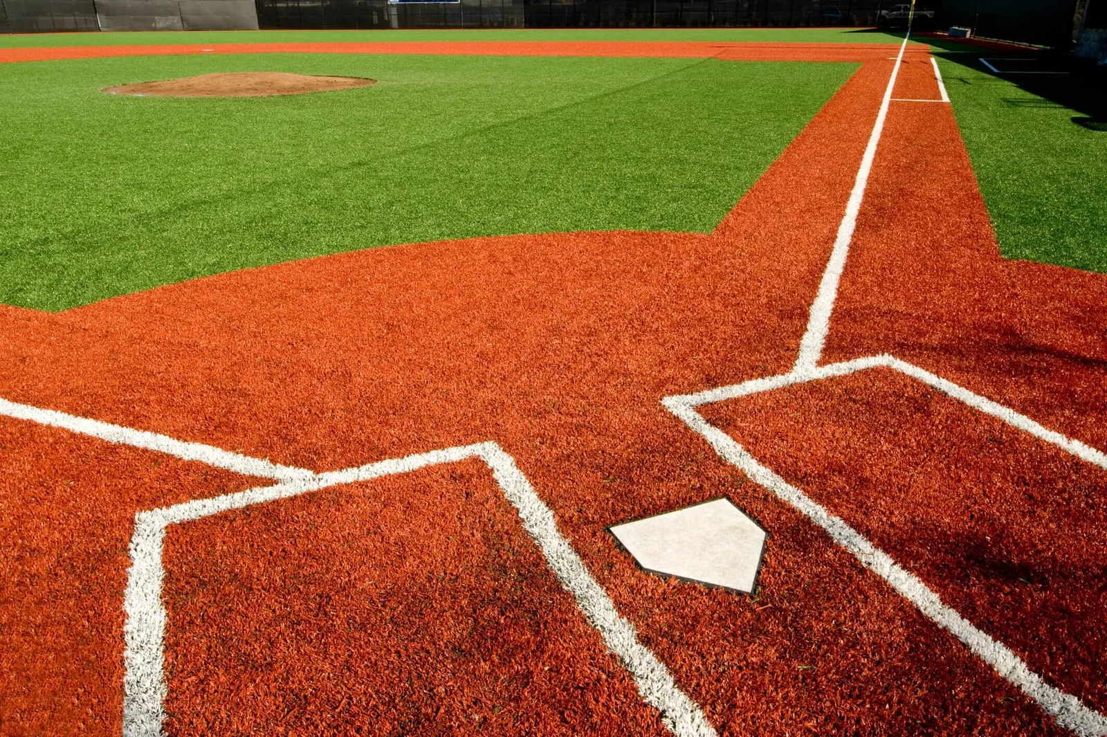 The baseball field showcases home plate and the surrounding red artificial turf, with white chalk lines marking the batter's box and foul line. The vibrant green synthetic turf in the outfield and the pitcher's mound are clearly visible in the distance.